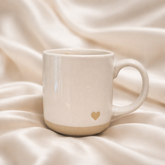 Minimal white ceramic mug with a small gold heart detail, styled on soft neutral satin in an elegant product photo